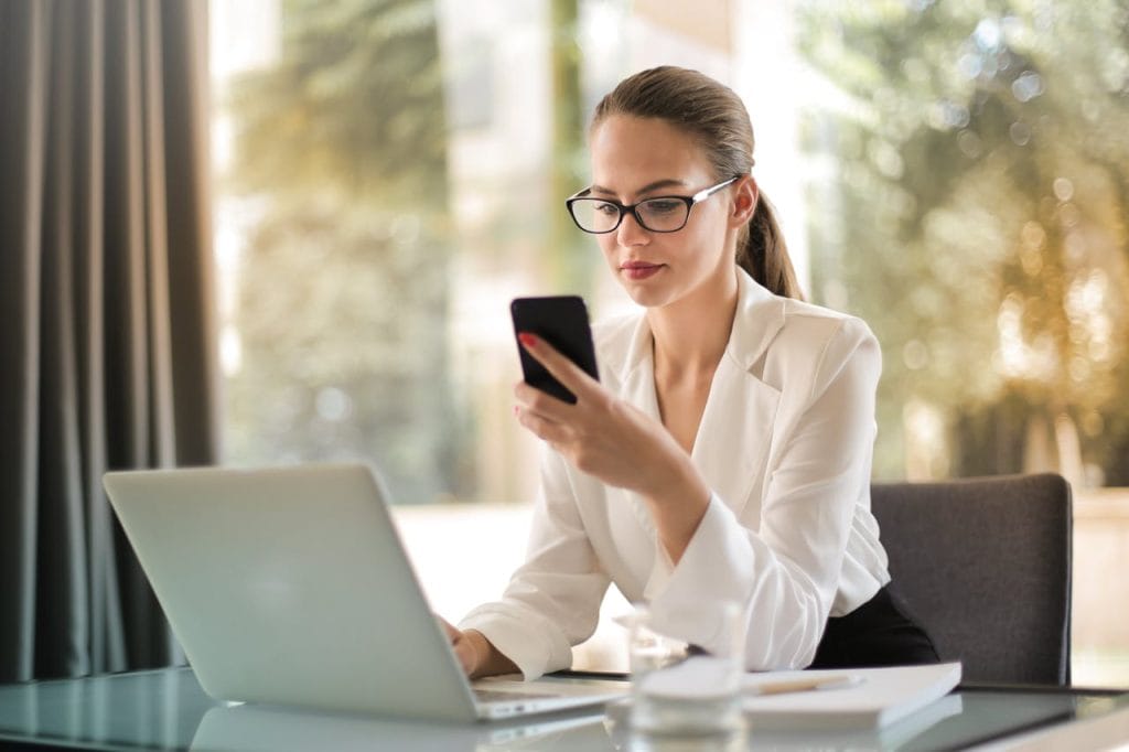 Professional woman using smartphone while working on laptop in modern office.