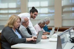 Senior adults use laptops in a classroom setting, guided by a young instructor.