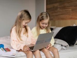 Two young girls engaged in online learning on a laptop while sitting on a cozy bed.